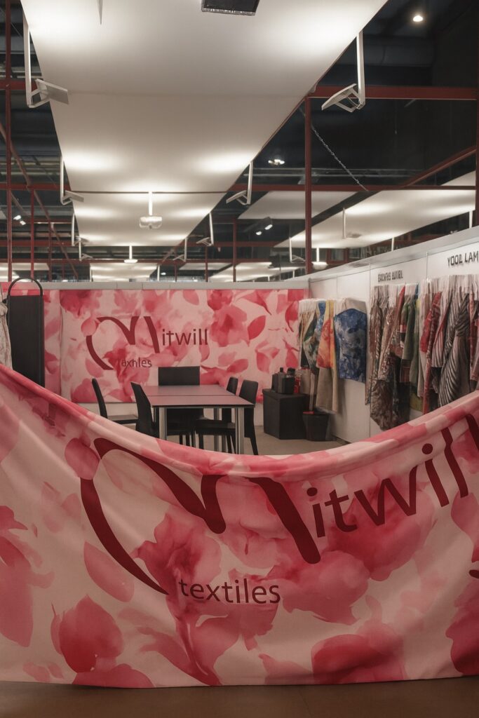 Mitwill Textiles booth at Milano Unica trade fair, with pink floral fabric and colorful samples displayed on the walls.